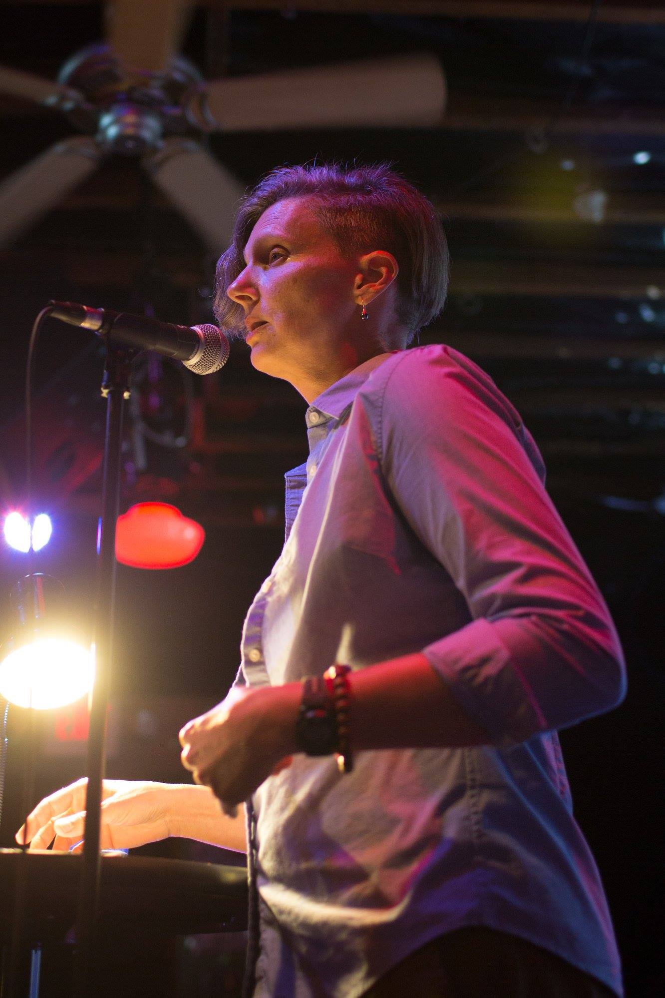 Photo of a middle-aged woman in front of a microphone in a darkened venue. The photo is taken from a low angle off to the side and close up. The woman has the hair on the left side of her head cropped short and is wearing a button-up collared shirt with a tank top underneath.