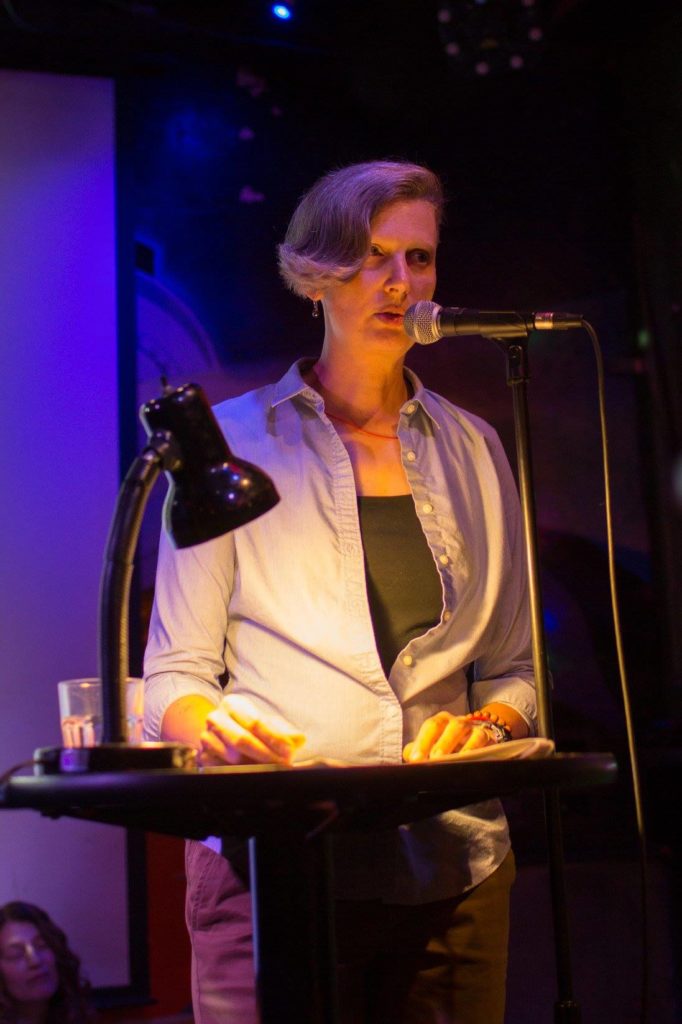 woman with short blonde hair standing at a microphone in a room with a darkened background