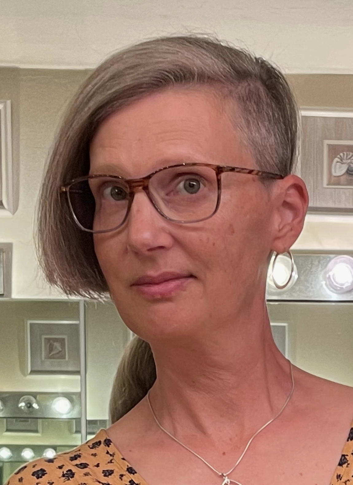 Head shot of a woman with cropped blonde hair on one side of her head and wearing glasses.